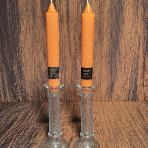 May include: Two orange taper candles in clear glass candle holders. The candles have a textured surface and a black label near the base. The candle holders have a detailed floral design. The candles are lit.