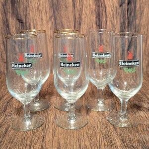 May include: Six clear beer glasses with gold rims. Each glass has the Heineken logo in a black rectangle with a red windmill and green accents. The glasses are on a brown surface.