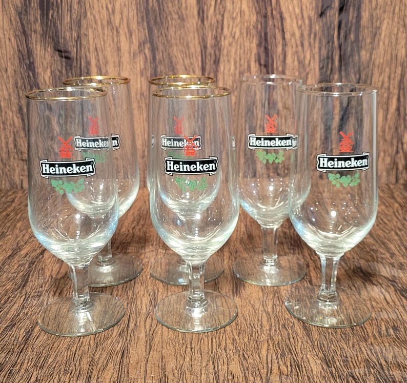 May include: Six clear beer glasses with gold rims. Each glass has the Heineken logo in a black rectangle with a red windmill and green accents. The glasses are on a brown surface.