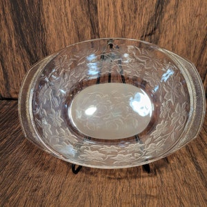 May include: A clear glass oval serving bowl with a floral pattern etched into the glass. The bowl has a raised rim and is sitting on a wooden surface.