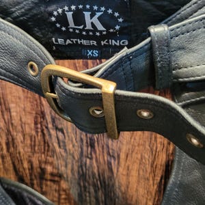 May include: Close-up of a black leather garment with a buckle and the text "LEATHER KING". The garment has a belt with a gold-coloured buckle and the size "XS" is visible.