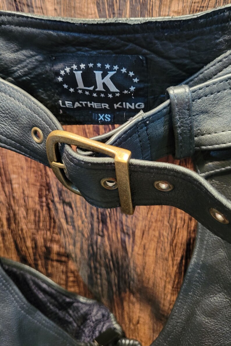 May include: Close-up of a black leather garment with a buckle and the text "LEATHER KING". The garment has a belt with a gold-coloured buckle and the size "XS" is visible.