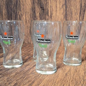 May include: Four clear glass Heineken beer glasses. Each glass has the Heineken logo with a red star and green accents. The glasses are empty and sit on a brown wooden surface.