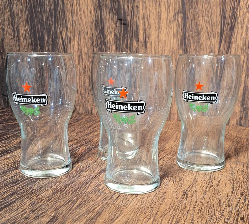 May include: Four clear glass Heineken beer glasses. Each glass has the Heineken logo with a red star and green accents. The glasses are empty and sit on a brown wooden surface.