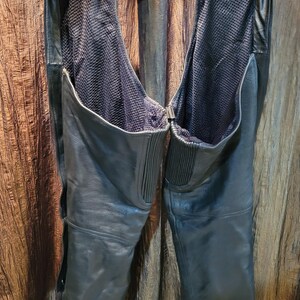 May include: Black leather motorcycle chaps with a mesh lining. The chaps have a belt and zip closure. The leather is smooth and the chaps are hanging from a metal hanger.