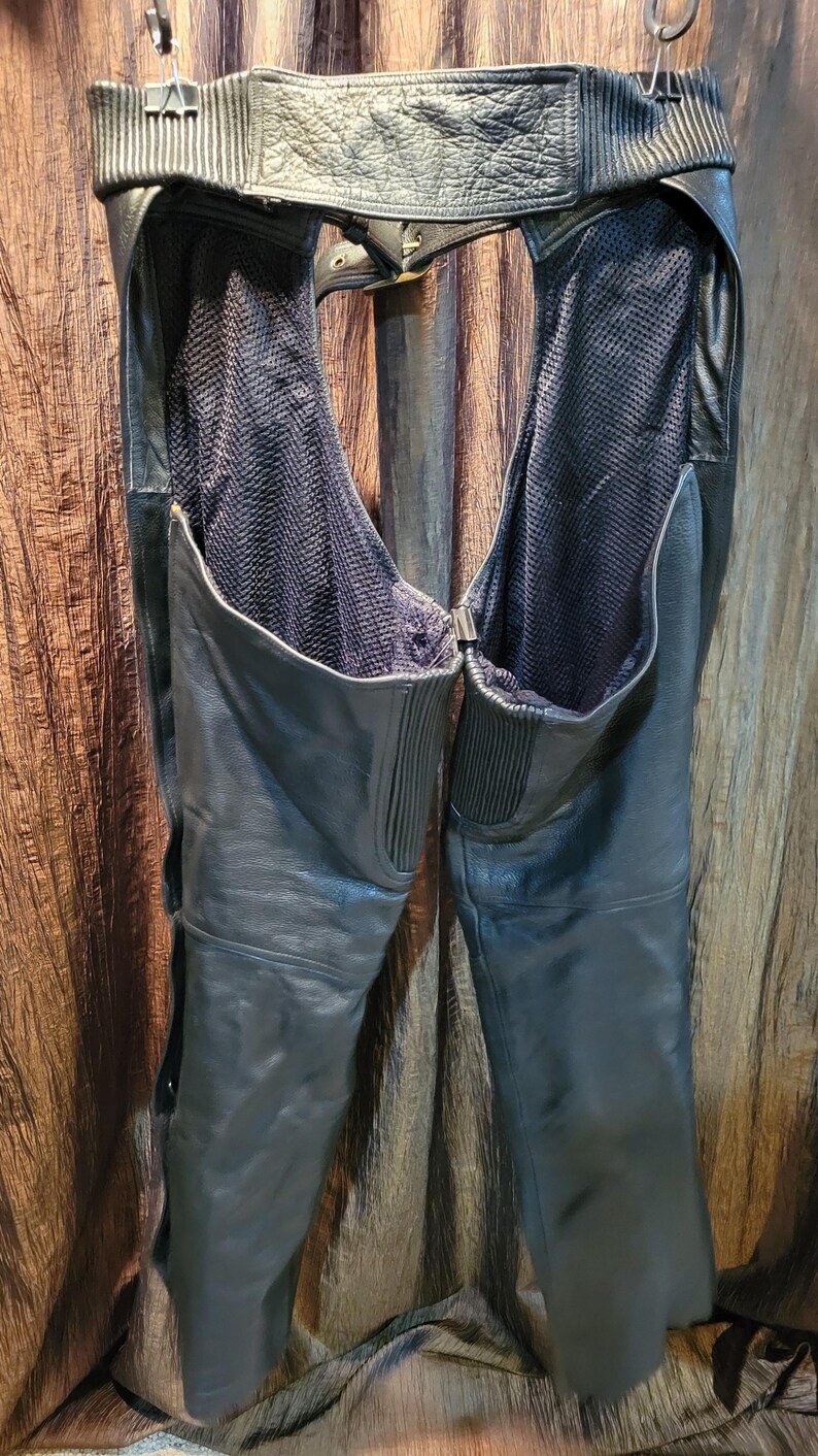 May include: Black leather motorcycle chaps with a mesh lining. The chaps have a belt and zip closure. The leather is smooth and the chaps are hanging from a metal hanger.