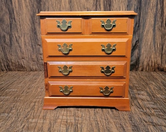 May include: A wooden jewelry box with five drawers and brass hardware. The box is a light brown color and has a simple design.