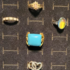 May include: A collection of rings displayed on a black surface. The rings feature various designs, including a gold band with a pink stone, a silver band with a turquoise stone, and a silver ring with a heart design.