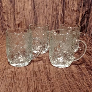 May include: Four clear glass mugs with a floral pattern. The mugs have a rounded shape and a handle.