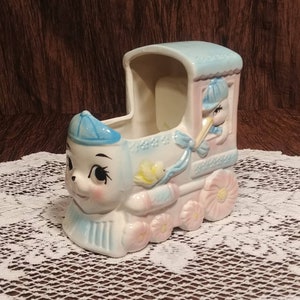 May include: A ceramic train planter with a blue and pink color scheme. The front of the train features a cartoon animal face with large eyes. The back of the train has a window with a cartoon stork inside. The train has wheels with pink flowers on them.