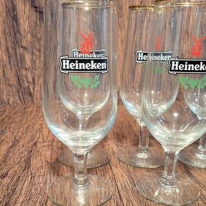May include: A set of clear, stemmed beer glasses with gold rims. Each glass features the Heineken logo in black and white with a red windmill and green accents. The glasses are arranged on a wooden surface.