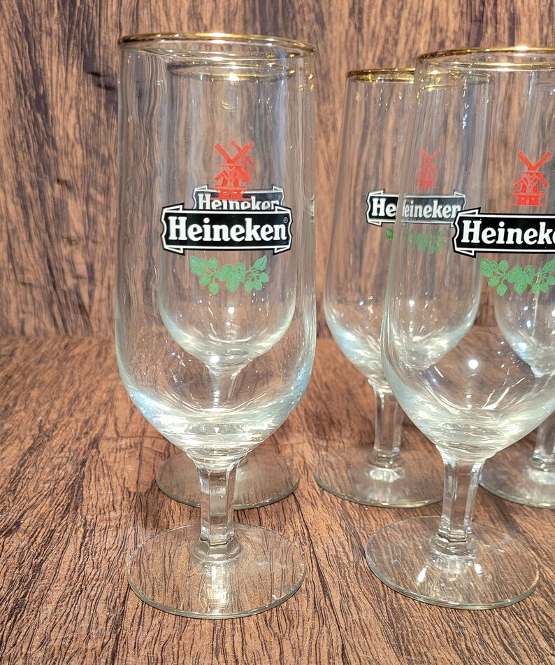 May include: A set of clear, stemmed beer glasses with gold rims. Each glass features the Heineken logo in black and white with a red windmill and green accents. The glasses are arranged on a wooden surface.