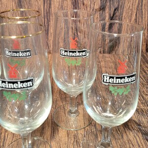May include: Several clear, stemmed beer glasses with the Heineken logo. The logo features a red windmill and green leaves. Some glasses have a gold rim. The glasses are on a brown wooden surface.