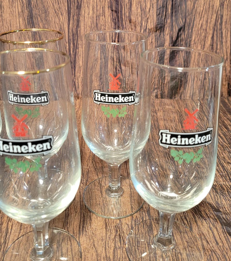 May include: Several clear, stemmed beer glasses with the Heineken logo. The logo features a red windmill and green leaves. Some glasses have a gold rim. The glasses are on a brown wooden surface.