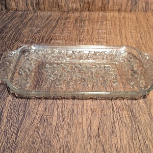 May include: A clear, rectangular glass baking dish with decorative floral patterns. The dish has handles on each end and is designed for oven use. The glass is transparent, allowing visibility of the contents. The dish is approximately 12 inches long.