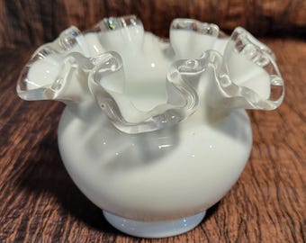 May include: A white glass vase with a ruffled rim. The vase has a round body and a clear, wavy edge. The vase is sitting on a brown surface.