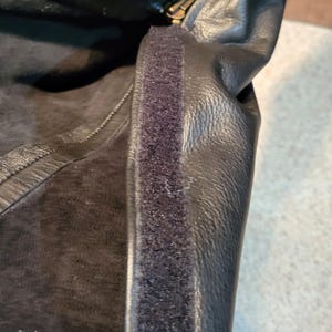 May include: Close-up of a black leather item with a zip and a strip of black velcro. The leather has a textured appearance. The zip is gold-coloured.