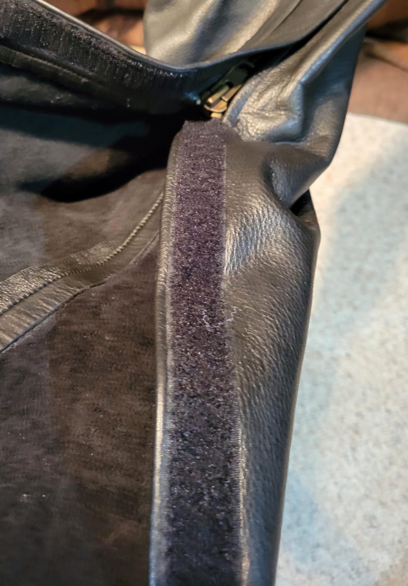 May include: Close-up of a black leather item with a zip and a strip of black velcro. The leather has a textured appearance. The zip is gold-coloured.