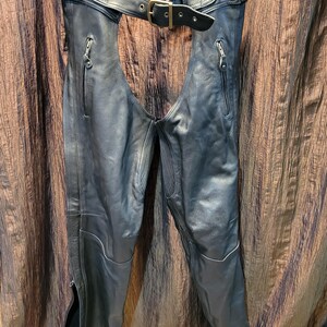 May include: Black leather motorcycle chaps with a belt and buckle closure. The chaps have zippered pockets and zippers along the outer leg seams. The waist area features a diamond-stitched design. The brand logo "LK" is visible on the back of the belt.