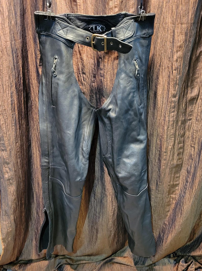 May include: Black leather motorcycle chaps with a belt and buckle closure. The chaps have zippered pockets and zippers along the outer leg seams. The waist area features a diamond-stitched design. The brand logo "LK" is visible on the back of the belt.