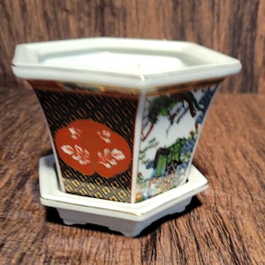 May include: A hexagonal ceramic planter with a matching saucer, featuring a black and gold geometric pattern, a prominent orange floral motif, and a detailed landscape illustration with green and blue accents, finished with a gold-trimmed rim.
