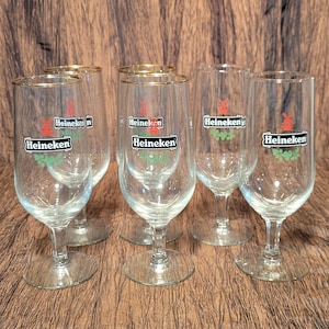 May include: Six clear beer glasses with gold rims. Each glass has the Heineken logo in red, green, and white. The glasses are on a brown surface.