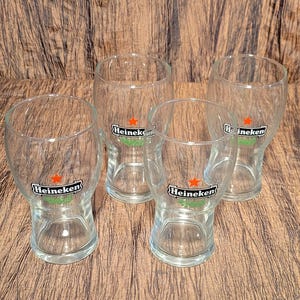 May include: Four clear beer glasses with the Heineken logo, featuring a green banner and a red star. The glasses are empty and arranged on a wooden surface.