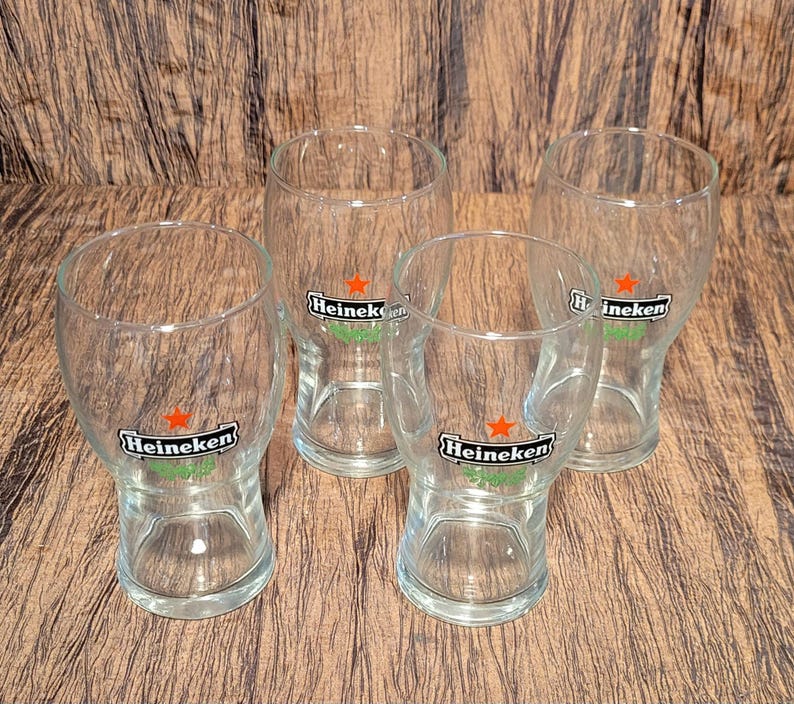 May include: Four clear beer glasses with the Heineken logo, featuring a green banner and a red star. The glasses are empty and arranged on a wooden surface.