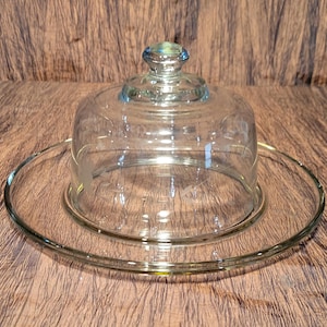 May include: A clear glass cloche with a round base and a domed lid. The cloche is designed to cover and protect food.
