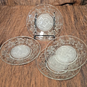 May include: Clear glass plates with a frosted floral design. The plates have a central floral motif and a border of leaves. One plate is on a black stand, and the others are stacked. The plates are on a brown surface.