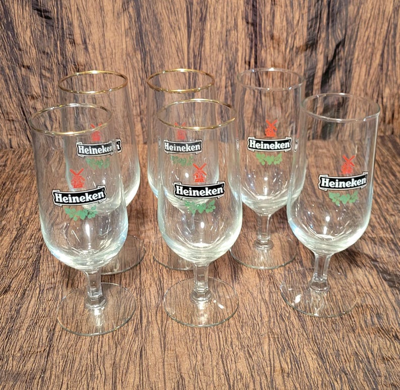 May include: Set of six clear stemmed beer glasses. Each glass has a gold rim and the Heineken logo in red, green, and white. The glasses are arranged on a brown wooden surface.