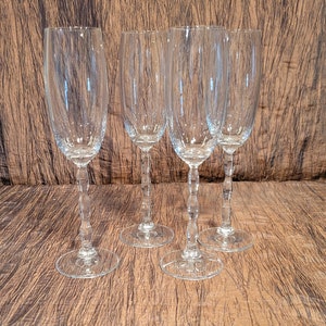 May include: Four clear glass champagne flutes on a brown textured surface. The flutes are tall and slender with a rounded bowl and a long stem.