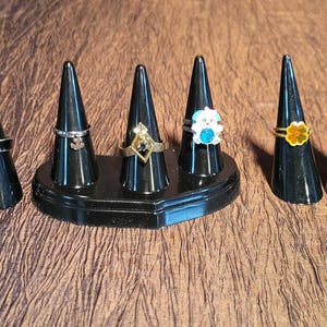 May include: A selection of rings presented on black, cone-shaped stands. The rings showcase diverse designs, such as a black band, an anchor charm, a geometric shape with a dark stone, a white flower with a blue centre, a yellow flower, and a crescent moon.
