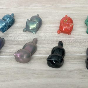 May include: A collection of ten small resin cat figurines in various colors, including black, blue, green, red, and purple. The cats are all in a curled-up position, with their tails tucked under their bodies.