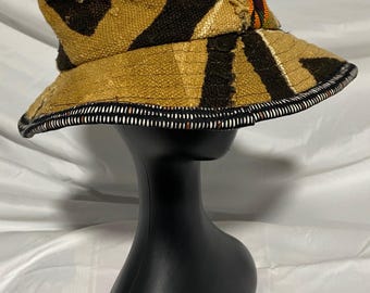 May include: A tan and black patterned bucket hat with a wide brim. The hat features a decorative band of white and black along the brim's edge. The hat is adorned with small white shells and colorful beads.