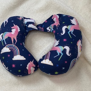May include: A blue travel pillow shaped like a horseshoe with a unicorn pattern. The pillow is covered in a navy blue fabric with pink unicorns, rainbows, and stars.
