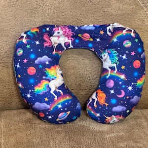 May include: A U-shaped travel pillow featuring a dark blue background with a repeating pattern of white unicorns, colorful rainbows, bright stars, purple clouds, and various planets in shades of red, orange, and blue across the entire fabric surface.