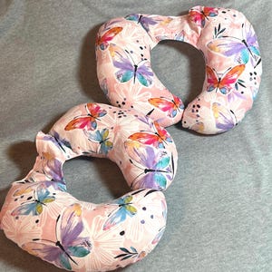 May include: Two pink and white donut-shaped pillows with a butterfly print. The pillows are made of soft fabric and have a comfortable shape.