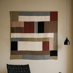 Puede incluir: Una escena de salón moderno con una silla de cuero negro tejido, un gran tapiz de pared geométrico en tonos neutros y dos jarrones decorativos. Una lámpara colgante negra cuelga del techo, lo que se suma a la estética contemporánea. El tapiz es en tonos marrones, grises y crema.