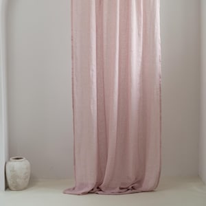 May include: A long, light pink curtain hangs in front of a white wall. The curtain is made of a sheer fabric and is gathered at the top. A white vase sits on the floor to the left of the curtain.