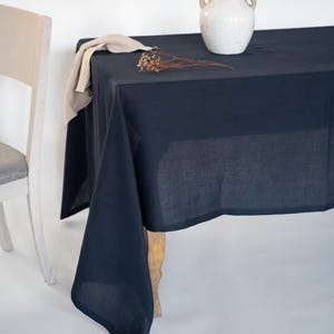 May include: A navy blue linen tablecloth draped over a wooden table. A white ceramic vase with dried flowers sits on the table. A beige linen napkin and dried flowers are also on the table.