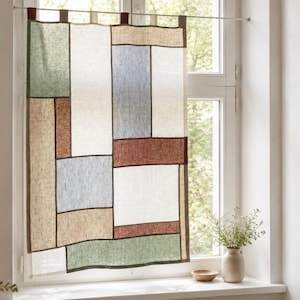Peut inclure: Un rideau de fenêtre rectangulaire présentant un motif patchwork de panneaux en tissu à texture de lin dans des tons sourds de vert sauge, terre cuite, bleu clair, crème et beige, séparés par des bordures marron foncé et suspendus à une fine tringle.