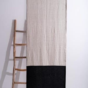 Puede incluir: Una cortina larga y rectangular de lino con un dise&ntilde;o bicolor. La parte superior es de color beige claro, mientras que la inferior es negra. Una escalera de madera se apoya en la pared a la izquierda de la cortina. La cortina cuelga de una barra de metal.