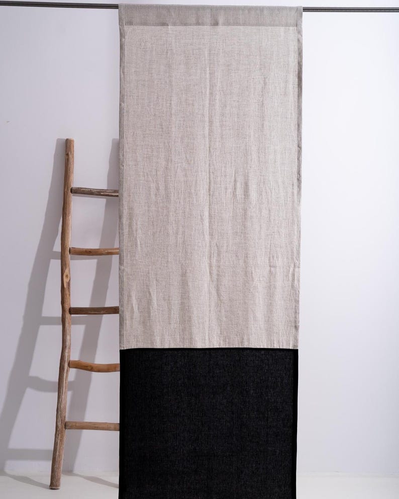 Puede incluir: Una cortina larga y rectangular de lino con un dise&ntilde;o bicolor. La parte superior es de color beige claro, mientras que la inferior es negra. Una escalera de madera se apoya en la pared a la izquierda de la cortina. La cortina cuelga de una barra de metal.