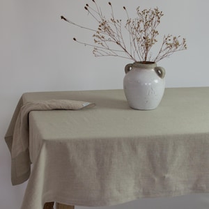 May include: A beige linen tablecloth draped over a wooden table with a white ceramic vase filled with dried flowers.