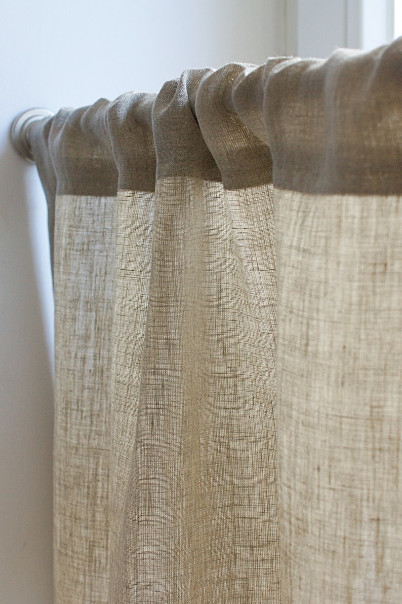 May include: Beige linen curtains hanging from a silver curtain pole. The curtains feature a textured weave and a darker beige band at the top. The fabric drapes in soft folds, creating a warm and inviting atmosphere.