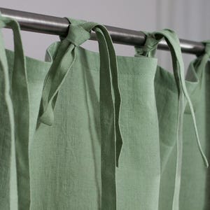 Puede incluir: Primer plano de cortinas de lino verde salvia colgadas de una barra de metal. Las cortinas están atadas con lazos de tela a juego. La tela tiene un aspecto texturizado y la estética general es sencilla y natural.