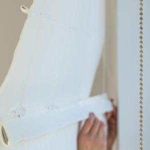 May include: A white, semi-sheer window shade is partially rolled up, revealing its layered texture. The shade is held in place by a series of clear plastic fasteners and strings. A decorative beaded strand hangs on the right side.
