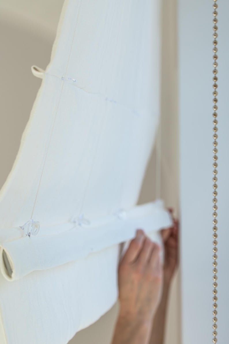 May include: A white, semi-sheer window shade is partially rolled up, revealing its layered texture. The shade is held in place by a series of clear plastic fasteners and strings. A decorative beaded strand hangs on the right side.