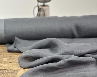 May include: A roll of dark gray linen fabric, partially unrolled, with a soft, textured appearance. The fabric is draped over a wooden surface, showcasing its natural drape and color. A decorative bird figurine is in the background.